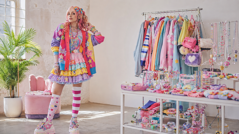 Woman wearing a colorful layered Decora Kei outfit with stacked accessories beside a bright rack of pastel Harajuku fashion.