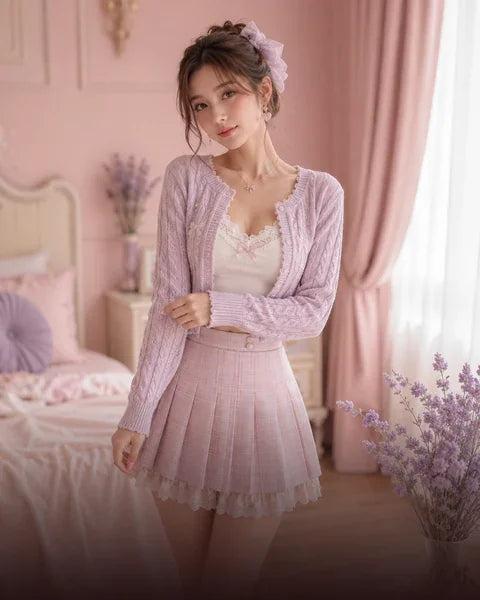 A young woman models a pastel pink three-piece outfit consisting of a cable-knit cardigan, a pleated mini skirt, and a lace-trimmed camisole top, set against a soft pink bedroom background.