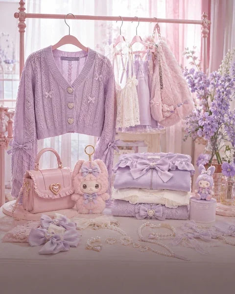 A pastel pink and lavender boutique display featuring a lavender knit cardigan sweater on a hanger, a pink handbag with a heart-shaped handle, a plush bunny toy, folded lace garments, and decorative accessories arranged on a table in front of a soft pink bed frame.